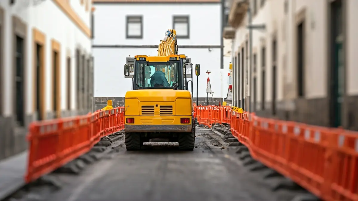 Imagen de obras en una calle estrecha con barreras de seguridad naranjas y maquinaria pesada.