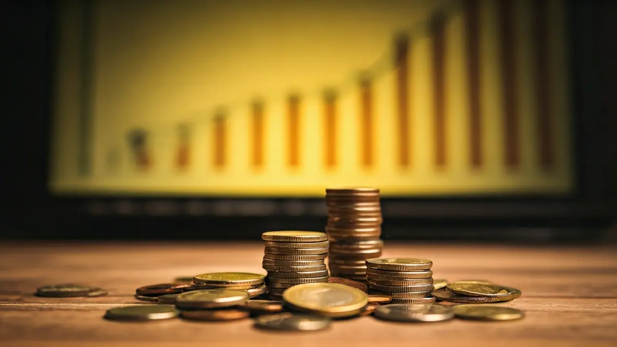 Imagen genérica de monedas y billetes sobre una mesa, con un gráfico financiero de fondo.