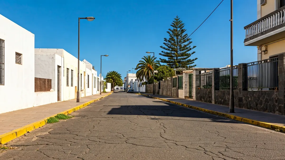 Imagen de una calle deteriorada en una urbanización, con asfalto agrietado y maleza.