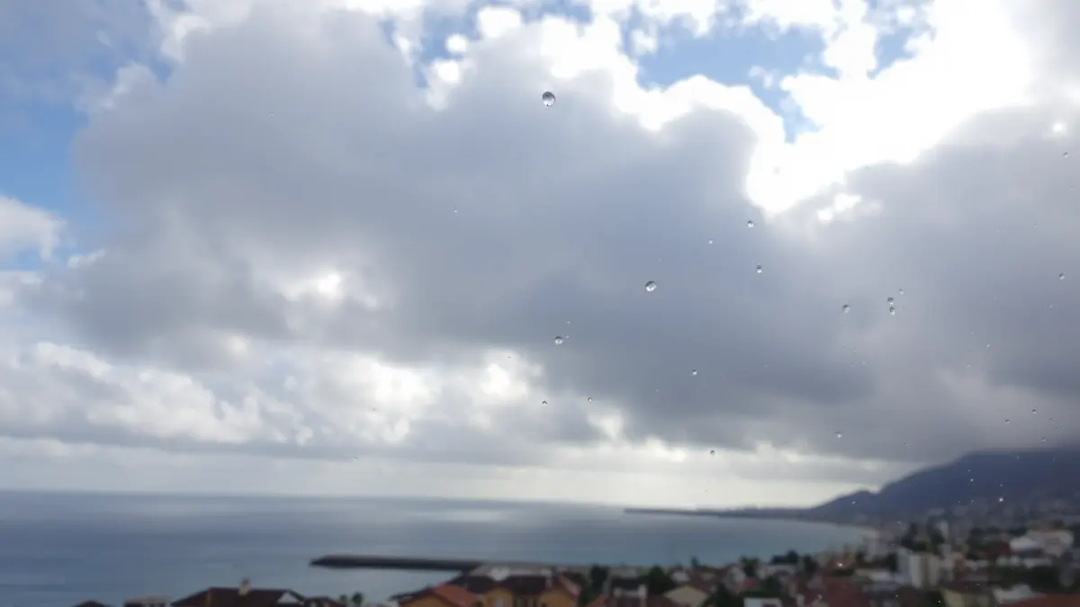 Imagen genérica de un cielo parcialmente nublado sobre una zona costera de Canarias, con algunos rayos de sol y gotas de lluvia en un cristal.
