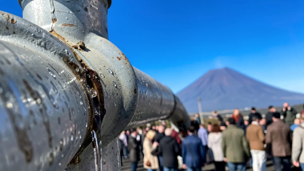 Primer plano de una tubería de agua con una ligera fuga, reflejando el cielo azul y un toque de paisaje volcánico al fondo.