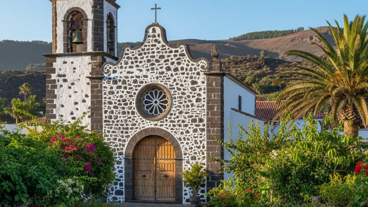 Imagen de una iglesia tradicional en un pueblo canario, con vegetación y montañas volcánicas.