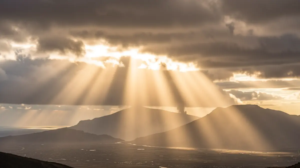 Cielo dramático sobre las Islas Canarias con intensos rayos de sol rompiendo a través de gruesas capas de polvo y nubes, indicando una ola de calor y calima. Paisaje volcánico al fondo.