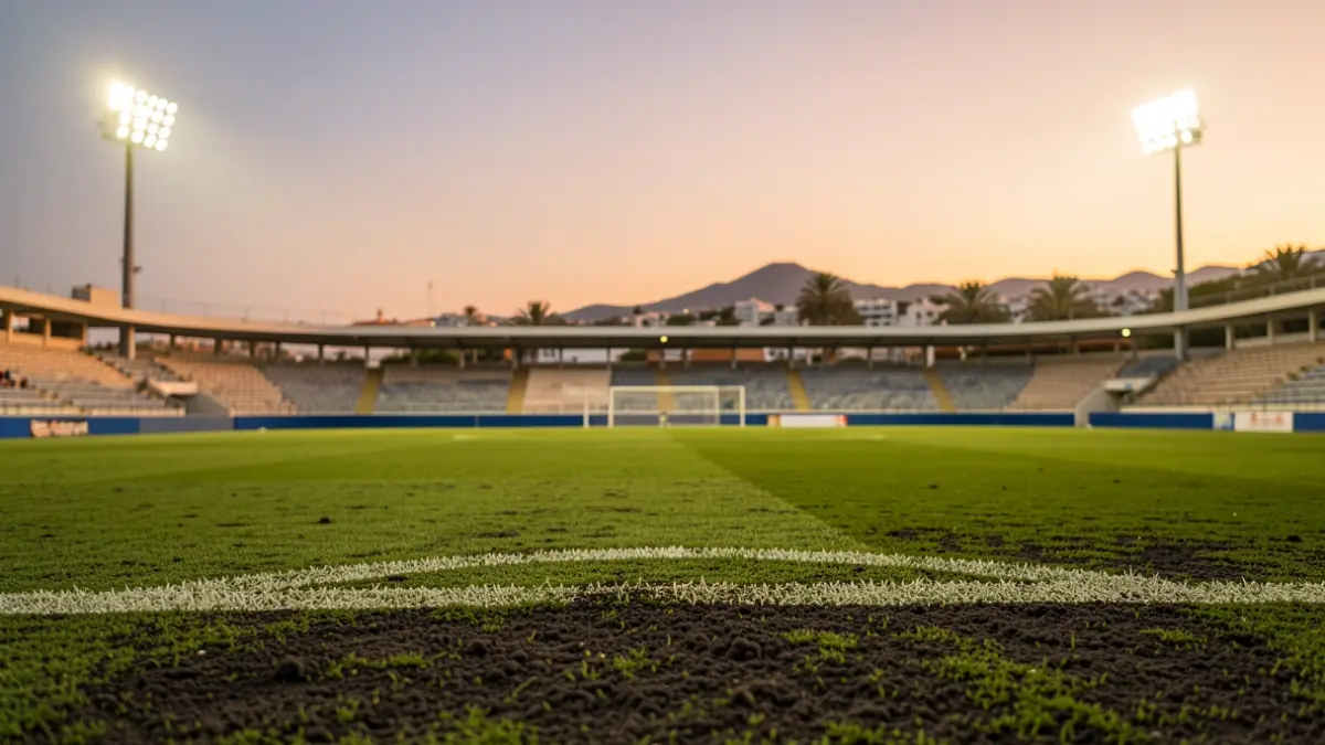 Football pitch in the Canary Islands with volcanic sand and green grass, stadium lights on, empty seats in the background.