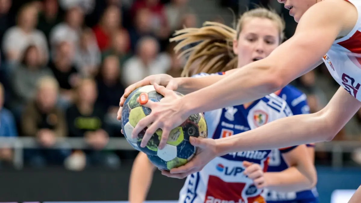 Primer plano de una jugadora de balonmano en acción, con el balón en las manos y el fondo de una cancha deportiva desenfocado.