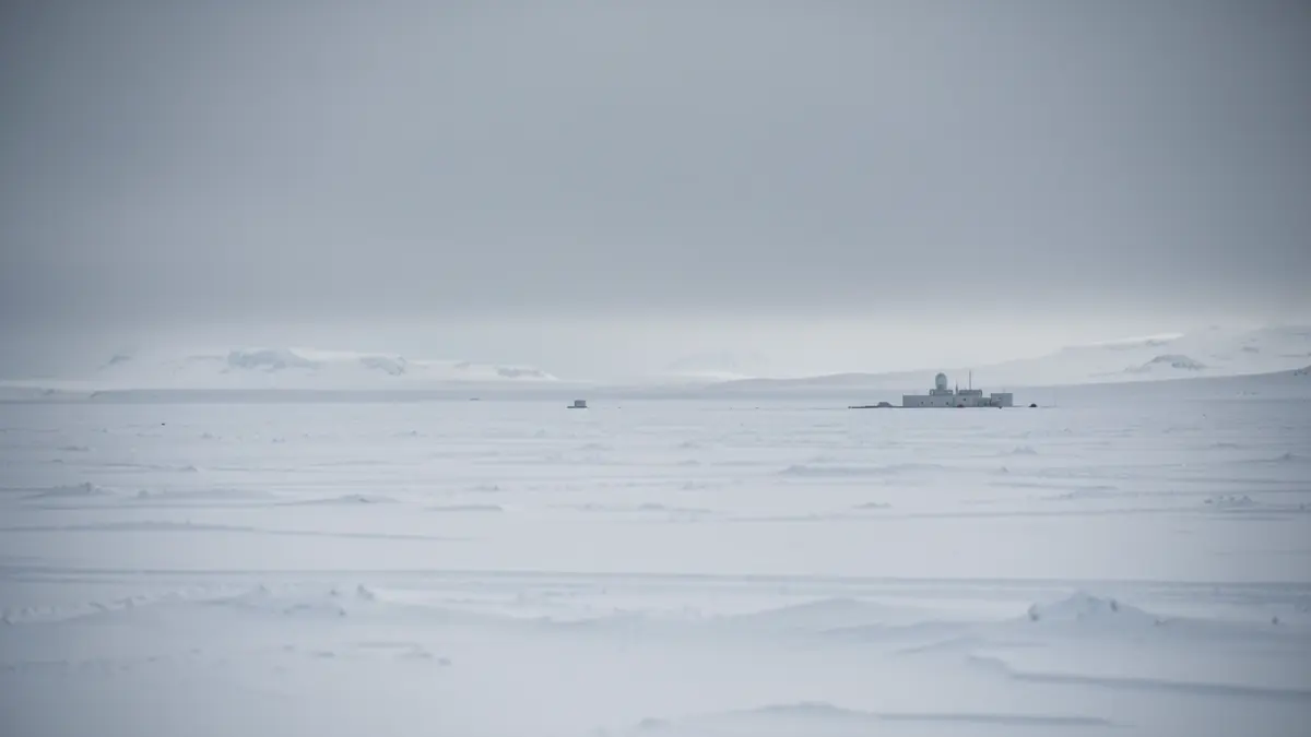Desolate Antarctic landscape with snow and a grey sky, showing a scientific base in the distance.