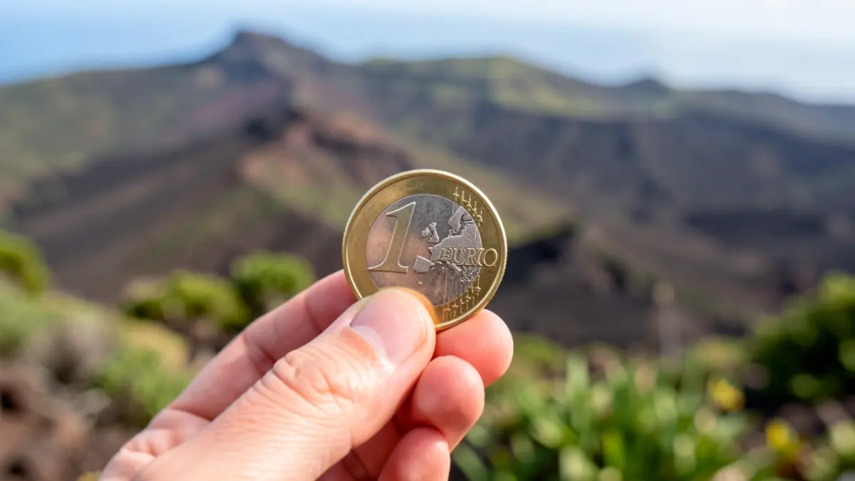 Primer plano de una mano sosteniendo una moneda de euro con un paisaje de Canarias al fondo.