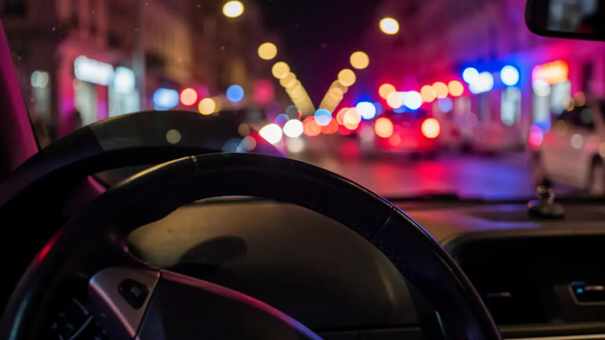 Primer plano del volante de un coche con el fondo borroso de una calle de noche, iluminado por luces de ciudad y de vehículos de emergencia.