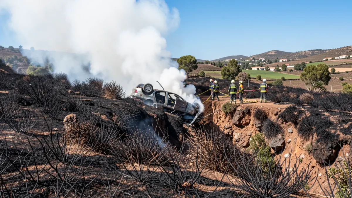 Imagen de un coche siniestrado en un barranco en Gáldar, Gran Canaria, con humo y bomberos actuando.