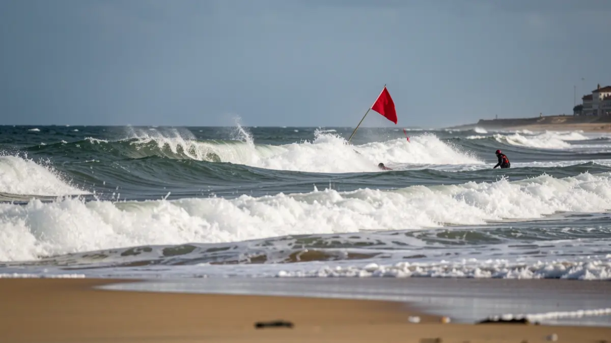 Imagen genérica de olas fuertes rompiendo en una playa con bandera roja ondeando, indicando peligro. Un socorrista en el agua se acerca a una persona.